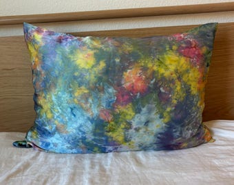Puede incluir: Un cojín cuadrado con un vibrante estampado tie-dye. El cojín presenta una mezcla de colores, incluyendo azul, amarillo, rojo y morado, creando un efecto acuarela. El cojín está colocado sobre una sábana blanca, con un cabecero de madera en el fondo.