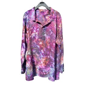 May include: A long-sleeved shirt with a vibrant tie-dye pattern in shades of purple, pink, and brown. The shirt features a collared neckline, a three-button placket, and buttoned cuffs. The garment is hanging on a black hanger against a white background.