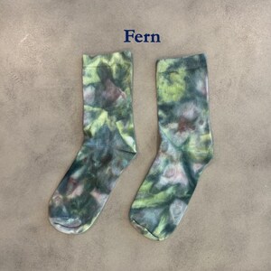 Puede incluir: Un par de calcetines de media ca&ntilde;a con un patr&oacute;n de te&ntilde;ido anudado en tonos verde oscuro, verde claro y marr&oacute;n apagado. La palabra Fern est&aacute; impresa en texto azul oscuro sobre los calcetines, que tienen un pu&ntilde;o acanalado y tejido est&aacute;ndar.
