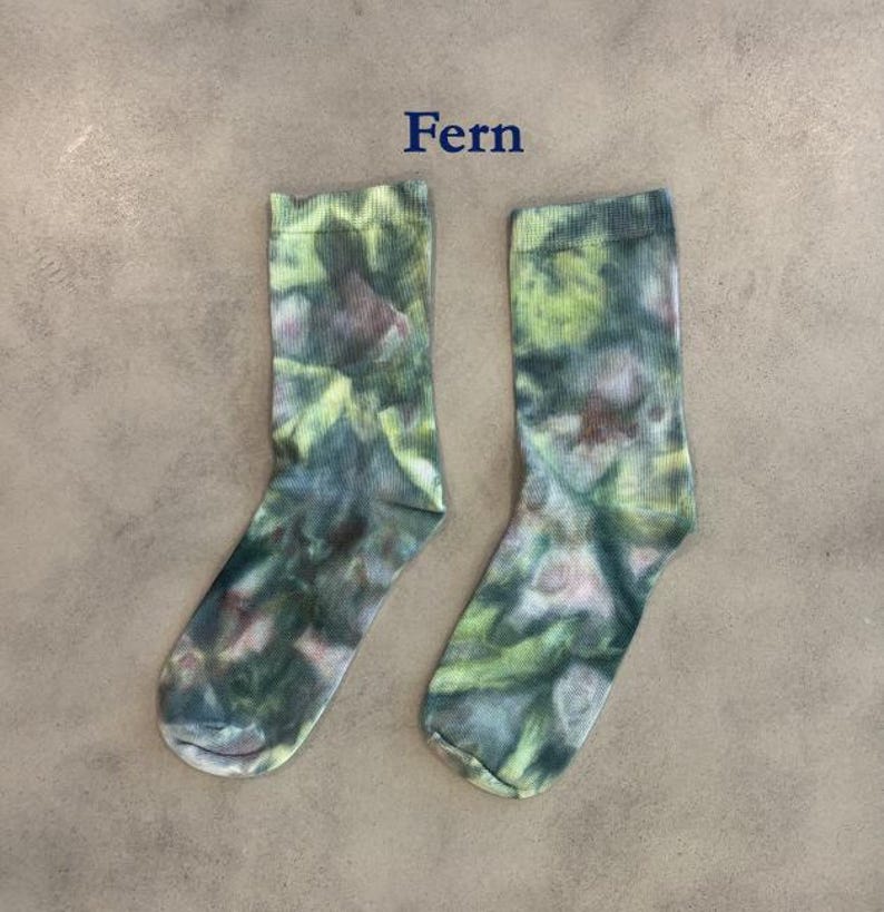 Puede incluir: Un par de calcetines de media ca&ntilde;a con un patr&oacute;n de te&ntilde;ido anudado en tonos verde oscuro, verde claro y marr&oacute;n apagado. La palabra Fern est&aacute; impresa en texto azul oscuro sobre los calcetines, que tienen un pu&ntilde;o acanalado y tejido est&aacute;ndar.
