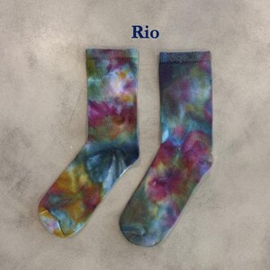 Puede incluir: Un par de calcetines de media ca&ntilde;a con un patr&oacute;n tie-dye multicolor con remolinos en magenta, verde azulado, amarillo y azul oscuro. La palabra Rio est&aacute; impresa en azul oscuro sobre los calcetines, que tienen un pu&ntilde;o acanalado y textura suave.