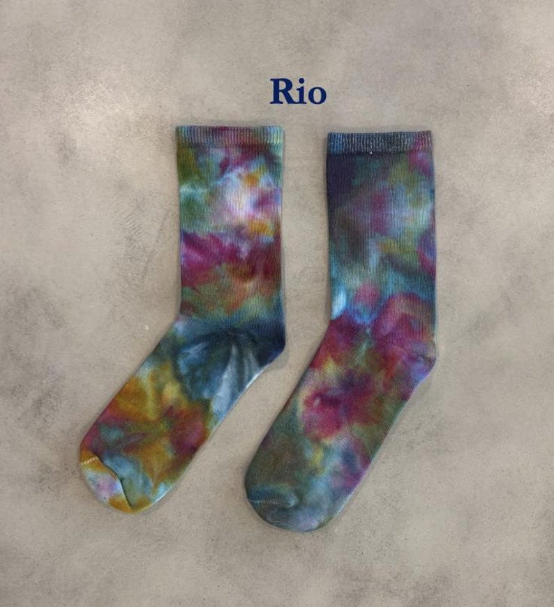 Puede incluir: Un par de calcetines de media ca&ntilde;a con un patr&oacute;n tie-dye multicolor con remolinos en magenta, verde azulado, amarillo y azul oscuro. La palabra Rio est&aacute; impresa en azul oscuro sobre los calcetines, que tienen un pu&ntilde;o acanalado y textura suave.
