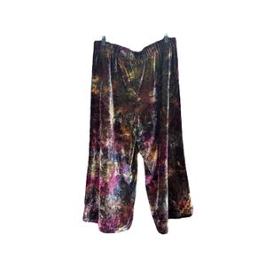 K&ouml;nnte beinhalten: Palazzo-Hose aus Samt mit Batikmuster. Die Hose hat ein weites Bein und einen elastischen Bund. Die Farben umfassen Braunt&ouml;ne, Lila, Gelb und Blau. Die Hose h&auml;ngt an einem schwarzen Kleiderb&uuml;gel vor wei&szlig;em Hintergrund.