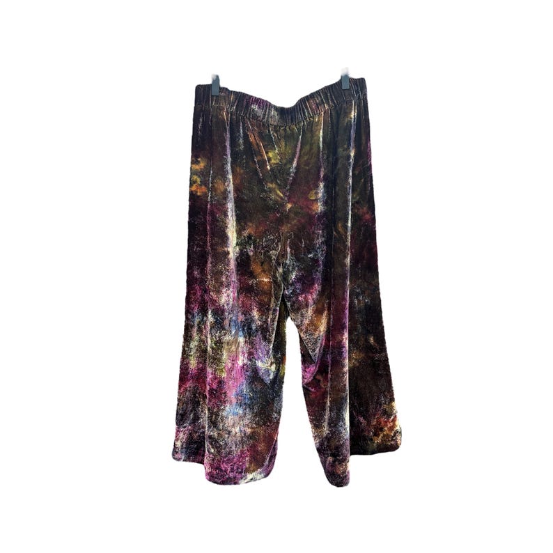 K&ouml;nnte beinhalten: Palazzo-Hose aus Samt mit Batikmuster. Die Hose hat ein weites Bein und einen elastischen Bund. Die Farben umfassen Braunt&ouml;ne, Lila, Gelb und Blau. Die Hose h&auml;ngt an einem schwarzen Kleiderb&uuml;gel vor wei&szlig;em Hintergrund.