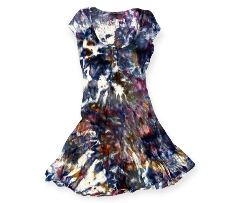 Pode incluir: Vestido tie-dye em tons de azul, branco, roxo e laranja. O vestido tem decote redondo, mangas curtas e saia rodada. Possui abotoamento frontal e uma pequena etiqueta perto da gola.