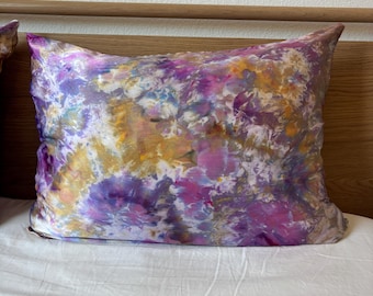 Puede incluir: Una funda de almohada rectangular con un estampado tie-dye en tonos morado, rosa, amarillo y blanco. La funda de almohada está sobre una sábana blanca, con un cabecero de madera en el fondo. Es un artículo decorativo.