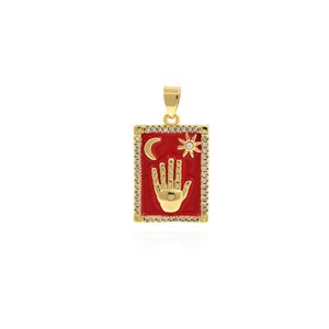Pode incluir: Pingente de ouro com um quadrado de esmalte vermelho que apresenta uma mão com uma lua crescente e um sol acima. O pingente é rodeado por strass transparentes.