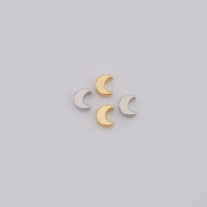 Peut inclure: Quatre petits charmes en forme de croissant de lune, deux dorés et deux argentés, sur un fond blanc.