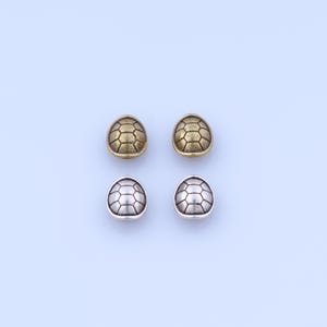 May include: Four metal turtle shell shaped beads. Two are gold-toned, and two are silver-toned. Each bead has a detailed, segmented pattern resembling a turtle's shell. The beads are arranged on a light blue background.