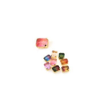 May include: A collection of colorful, rectangular gemstones with gold-colored settings. The gemstones display a range of hues, including pink, blue, green, and orange. The gems are arranged on a white background, showcasing their vibrant colors and intricate details.