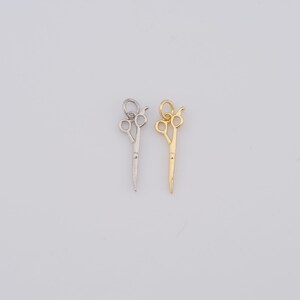 May include: Two pairs of scissors charms, one silver and one gold, hanging from a white background.