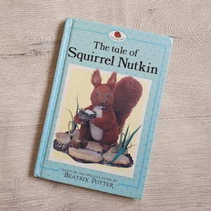 May include: A light blue hardcover book titled The Tale of Squirrel Nutkin by Beatrix Potter. The cover features a central illustration of a red squirrel holding a small basket, surrounded by rocks and grass, with a Ladybird logo at the top.