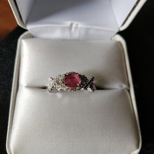 May include: A ring with a pink oval gemstone surrounded by smaller clear gemstones, set in a silver band. The ring is in a white ring box.