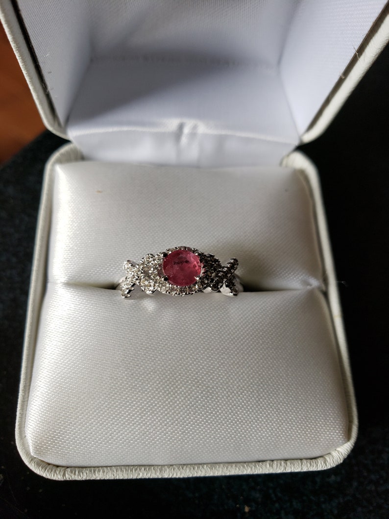 May include: A ring with a pink oval gemstone surrounded by smaller clear gemstones, set in a silver band. The ring is in a white ring box.