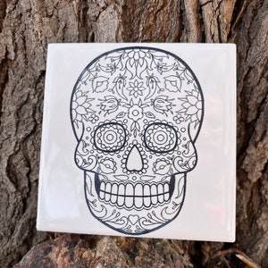 May include: Black and white sugar skull design on a white ceramic tile. The skull is decorated with intricate floral patterns.