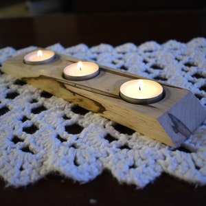 Puede incluir: Un portavelas de madera con tres velas de té encendidas. El portavelas está hecho de madera marrón claro con vetas marrones oscuras. Está sentado sobre un encaje blanco de ganchillo.