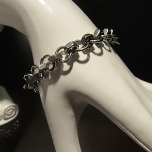 May include: A silver-toned bracelet with a unique design featuring hexagonal nut-shaped links and circular connectors. The bracelet is displayed on a white mannequin hand against a dark background, highlighting its metallic sheen and intricate construction.