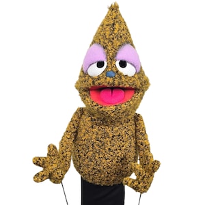 May include: A handmade puppet with a cone-shaped head and a textured, yellow and brown body. The puppet has large white eyes with purple eyelids, a blue nose, and a pink tongue. The puppet's mouth is open, and it has black clothing.