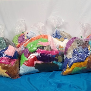 May include: Six clear plastic bags filled with colorful fabric scraps. The bags are arranged in a row on a blue surface.