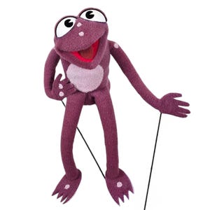 May include: A purple puppet frog with large white eyes and a red mouth. The frog has a light purple belly and spots. Black strings are attached to the frog's arms, suggesting it is a marionette.