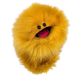 May include: A round, fluffy, mustard-yellow plush toy with long, soft synthetic fur. It features two small, round, black plastic eyes positioned above a wide, open mouth that reveals a bright pink, felt-like tongue inside the dark interior.