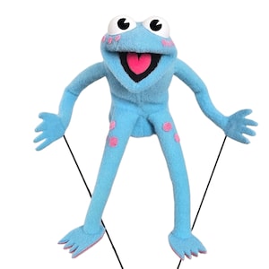 May include: A light blue puppet frog with large white eyes and a pink heart-shaped tongue. The frog has pink polka dots on its legs and cheeks. The puppet is suspended by black strings, suggesting it is a marionette toy.
