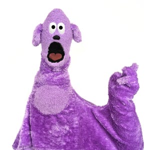 May include: A purple plush puppet featuring a long neck, two floppy ears, and a wide, open mouth with a dark interior. The puppet has two large white eyes with black pupils, a small black nose, and a circular patch on its chest.