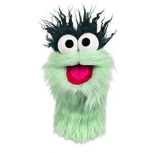 May include: A green and teal puppet with large white eyes, a pink tongue, and a green mustache. The puppet has a shaggy, furry texture and dark teal hair. The puppet is designed to be worn on the hand.