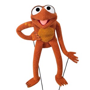May include: A plush orange frog puppet with large white-and-black eyes, a pink tongue, and a yellow belly. The puppet has articulated arms and legs, and is posed with one arm bent at the elbow. Black strings are attached for manipulation.