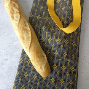 Puede incluir: Una bolsa de tela amarilla con un patrón geométrico en azul marino. La bolsa tiene dos asas largas amarillas y está llena de dos baguettes.