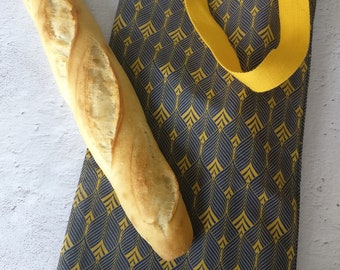 May include: A yellow tote bag with a geometric pattern in navy blue. The bag has two long yellow straps and is filled with two baguettes.