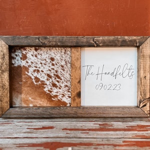 Op de afbeelding: Een rustiek houten frame met een gesplitst ontwerp. E&eacute;n kant heeft een bruin en wit koehuidpatroon, terwijl de andere kant een witte achtergrond heeft met de tekst "The Handfelts 090223" in zwarte cursieve letters.