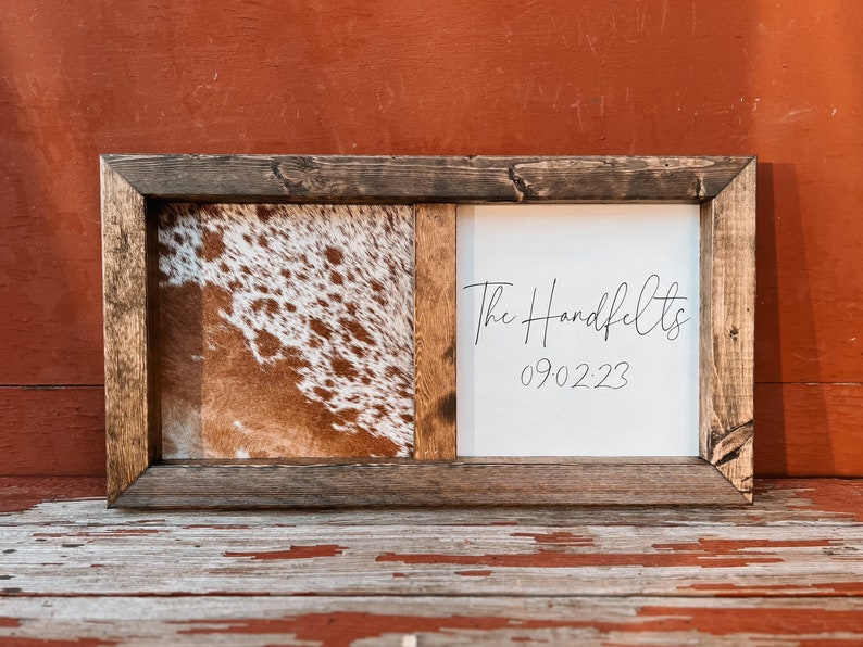 Op de afbeelding: Een rustiek houten frame met een gesplitst ontwerp. E&eacute;n kant heeft een bruin en wit koehuidpatroon, terwijl de andere kant een witte achtergrond heeft met de tekst "The Handfelts 090223" in zwarte cursieve letters.