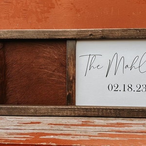 Op de afbeelding: Een houten ingelijst bord met een bruine lederen achtergrond en een wit canvas paneel met de tekst "The Mahlocks 02.18.23" in zwarte cursieve letters.