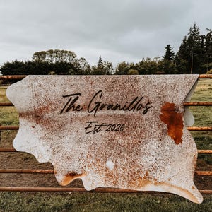 May include: A cowhide rug with a natural white and brown speckled pattern, featuring the black text "The Granillos" and "Est. 2026" printed in the center. The hide has an irregular, organic shape with distinct brown patches.