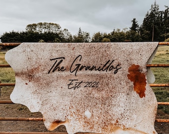 Op de afbeelding: Een koeienhuid met een natuurlijk wit en bruin gespikkeld patroon, voorzien van de zwarte tekst "The Granillos" en "Est. 2026" in het midden. De huid heeft een onregelmatige, organische vorm met opvallende bruine vlekken.