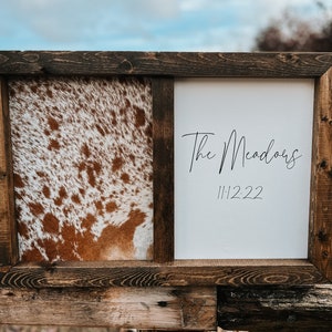 Op de afbeelding: Een bruin houten frame met een gesplitst ontwerp. E&eacute;n kant heeft een bruin en wit koehuidpatroon. De andere kant heeft een witte achtergrond met de tekst "The Meadows" en de datum "11.12.22" in zwart.