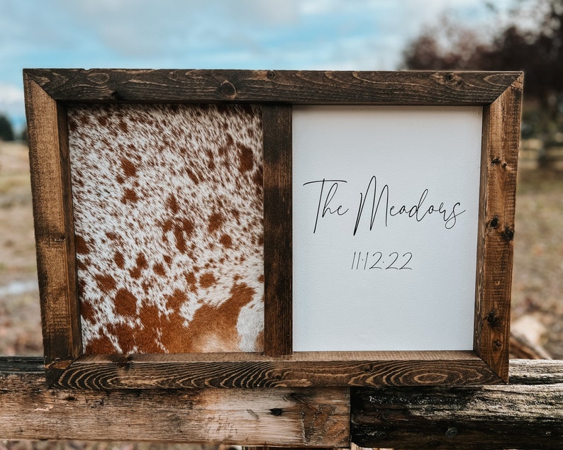 Op de afbeelding: Een bruin houten frame met een gesplitst ontwerp. E&eacute;n kant heeft een bruin en wit koehuidpatroon. De andere kant heeft een witte achtergrond met de tekst "The Meadows" en de datum "11.12.22" in zwart.