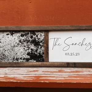 Op de afbeelding: Een houten ingelijst bord met een zwart-witte koehuidprint aan de linkerkant en een witte achtergrond met de tekst "The Sanchez 03.25.23" aan de rechterkant.