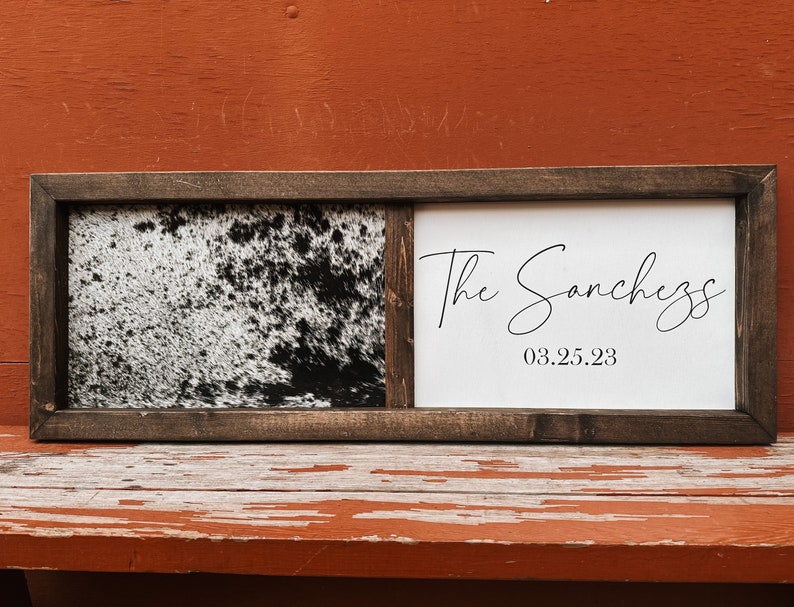 Op de afbeelding: Een houten ingelijst bord met een zwart-witte koehuidprint aan de linkerkant en een witte achtergrond met de tekst "The Sanchez 03.25.23" aan de rechterkant.
