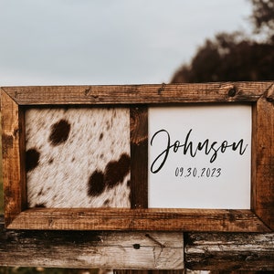 Op de afbeelding: Een houten ingelijst bord met een gesplitst ontwerp. E&eacute;n kant heeft een bruin en wit koehuidpatroon. De andere kant heeft een witte achtergrond met de tekst "Johnson 09.30.2023" in zwart.