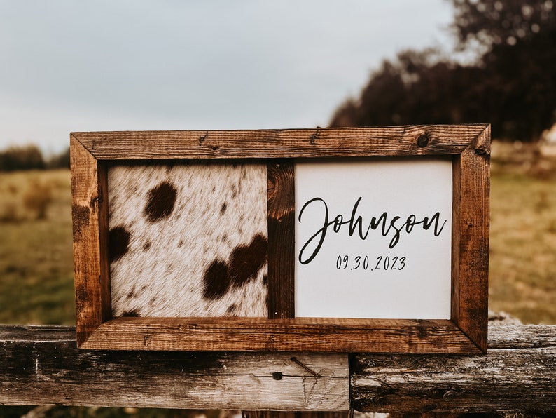 Op de afbeelding: Een houten ingelijst bord met een gesplitst ontwerp. E&eacute;n kant heeft een bruin en wit koehuidpatroon. De andere kant heeft een witte achtergrond met de tekst "Johnson 09.30.2023" in zwart.
