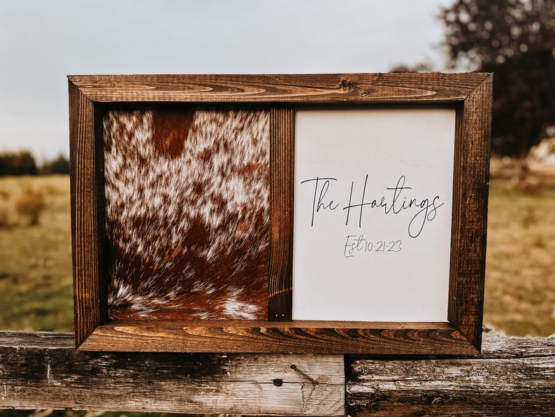 Op de afbeelding: Een houten frame met een gesplitst ontwerp. E&eacute;n kant heeft een bruin en wit koehuidpatroon. De andere kant heeft een witte achtergrond met de tekst "The Hartings Est. 10.21.23" in zwart.