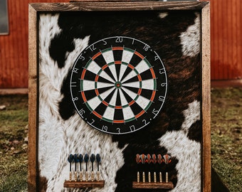 Op de afbeelding: Een dartbord met houten frame en een koeienhuid achtergrond. Het dartbord is zwart, wit en groen. Twee sets darts worden op houten houders weergegeven. De achtergrond is een mix van zwart-wit koeienhuid. Het frame is van donker hout.