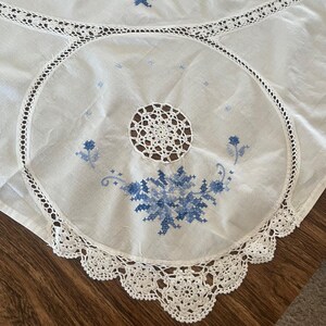 Puede incluir: Mantel blanco con intrincados bordados florales azules y un delicado borde de encaje de ganchillo. El centro presenta un dise&ntilde;o circular con un tapete de ganchillo. El mantel est&aacute; sobre una superficie de madera.