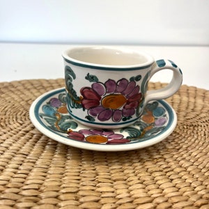 May include: A white ceramic teacup and saucer with a floral design. The cup and saucer are decorated with pink, purple, orange, and green flowers.