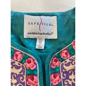 Może przedstawiać: Turkusowa bluzka z jedwabiu z kwiatowym nadrukiem i etykietą z napisem "Safe Silk by Adrianna Papell". Etykieta zawiera r&oacute;wnież informacje "100% jedwabiu" i "Wyprodukowano w Chinach".