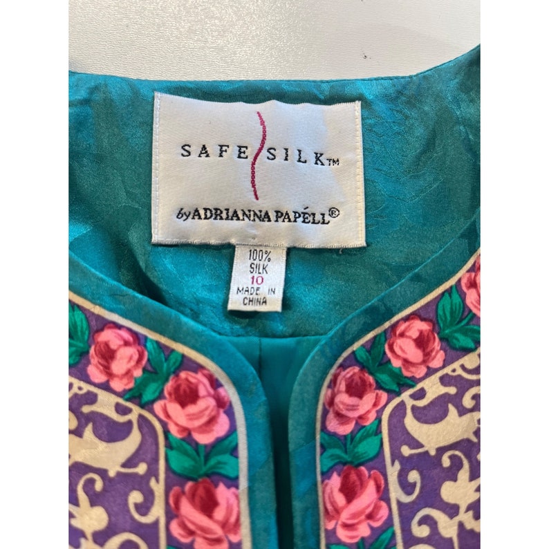 Może przedstawiać: Turkusowa bluzka z jedwabiu z kwiatowym nadrukiem i etykietą z napisem "Safe Silk by Adrianna Papell". Etykieta zawiera r&oacute;wnież informacje "100% jedwabiu" i "Wyprodukowano w Chinach".