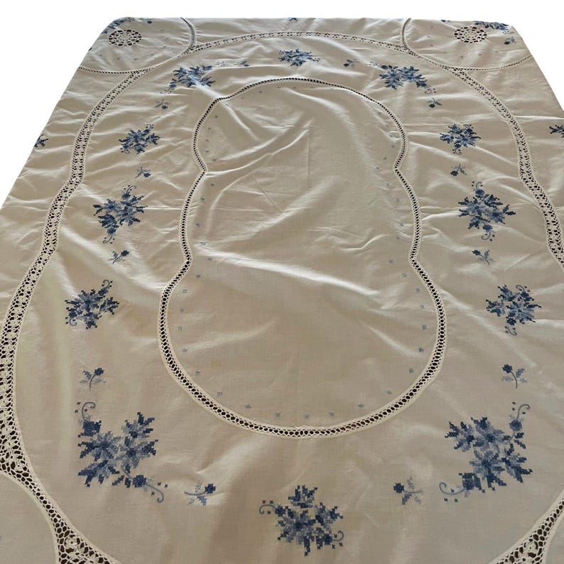 Puede incluir: Un mantel blanco con bordado floral azul. El mantel tiene un borde festoneado con un borde de ganchillo. El dise&ntilde;o floral se repite en la tela, creando un aspecto cl&aacute;sico y elegante.