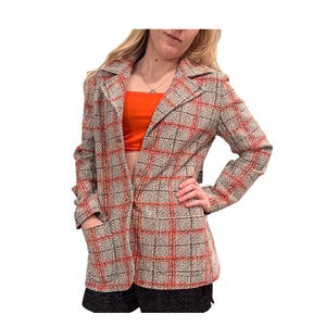 Puede incluir: Una chaqueta de traje de manga larga con un estampado de cuadros en tonos marrones, negros y naranjas. La chaqueta tiene un cuello de muesca, un cierre de botón único y dos bolsillos delanteros.